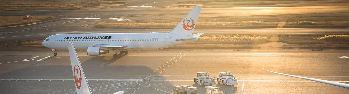 Japan Airlines aircraft on runway at golden-hour light with airport ground vehicles nearby.