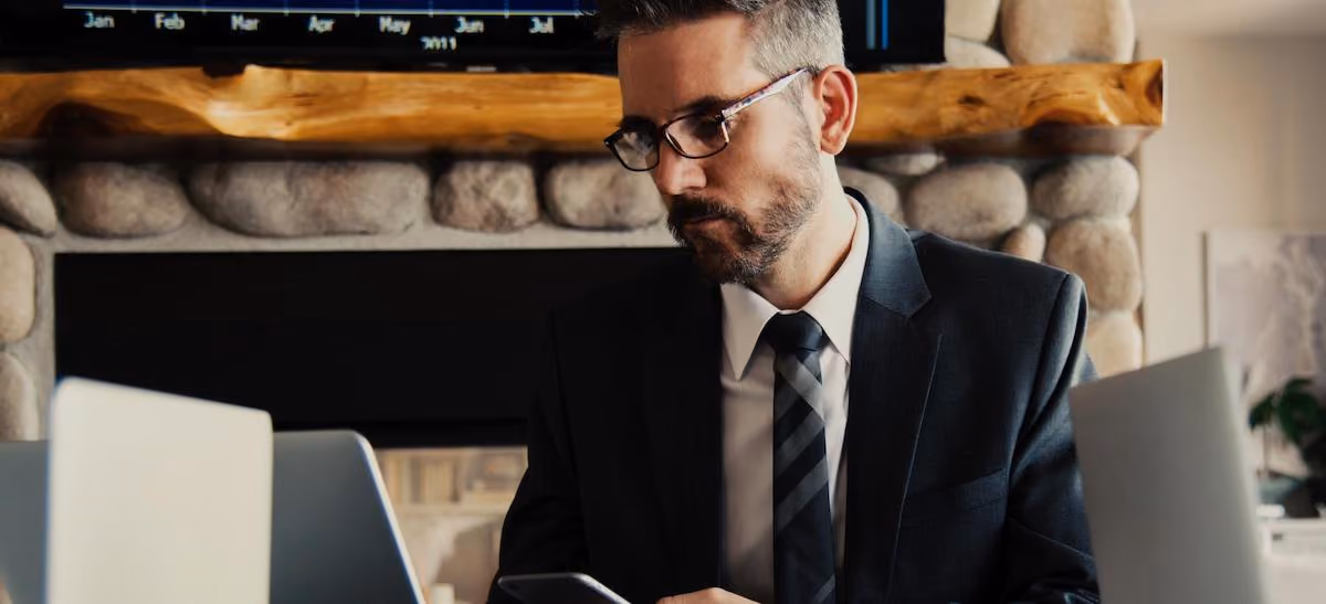 Man in suit and glasses looking at his phone with two laptops in front of him near stone fireplace.