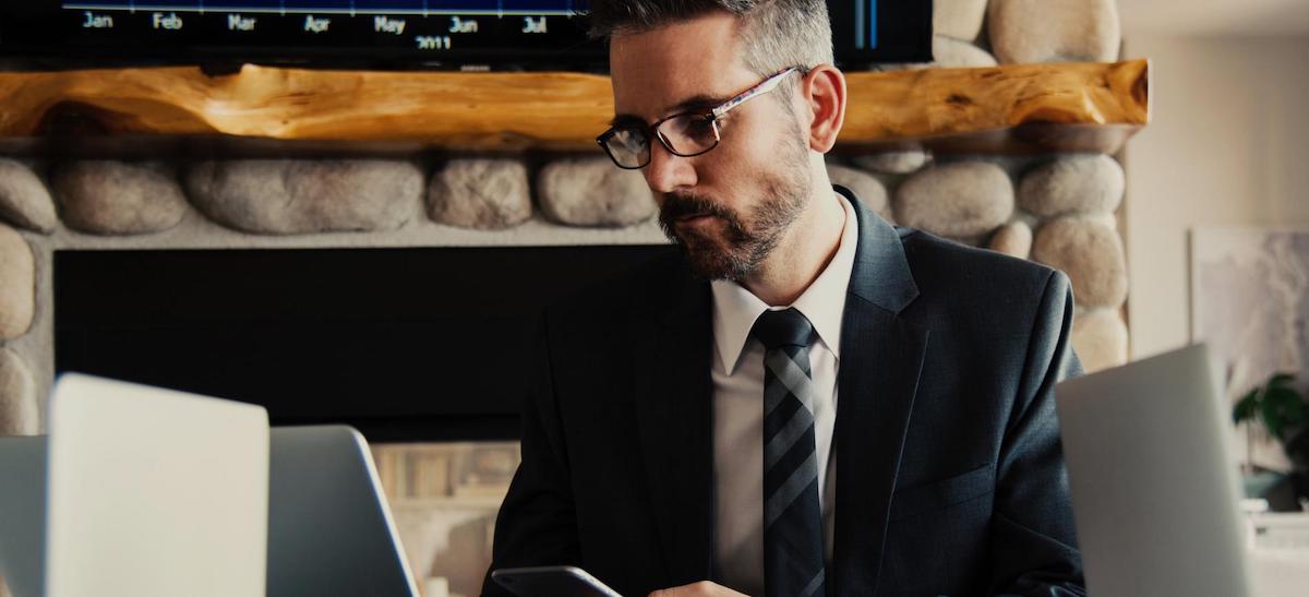 Man in suit and glasses looking at his phone with two laptops in front of him near stone fireplace.
