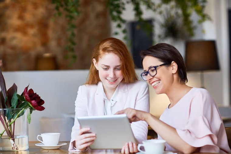 Two women smiling and looking at a tablet while sitting at a table with coffee cups and flowers.