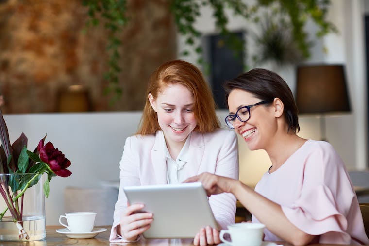 Two women smiling and looking at a tablet while sitting at a table with coffee cups and flowers.