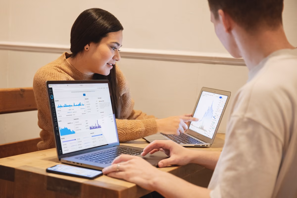 Two people at a table using laptops and phone, looking at financial charts and graphs on screens.