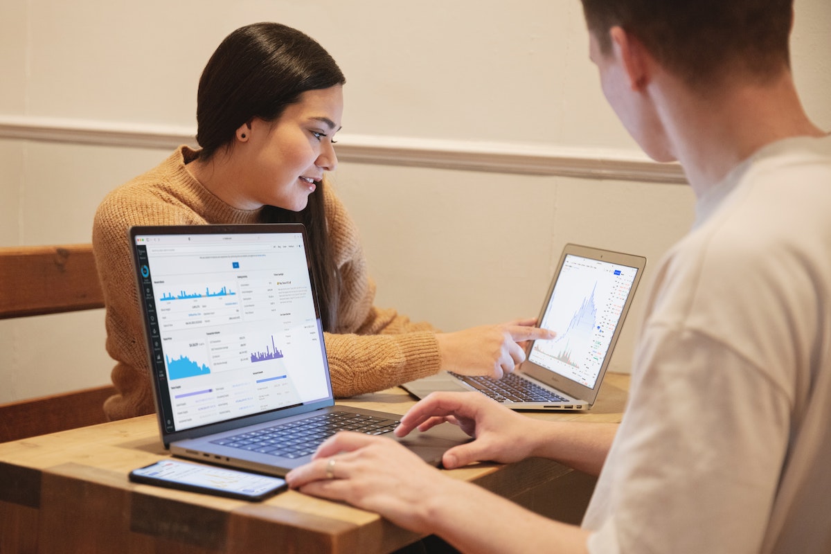 Two people at a table using laptops and phone, looking at financial charts and graphs on screens.
