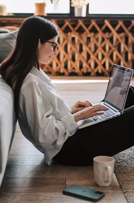 Woman with glasses sitting on floor leaning on sofa, typing on laptop with phone and mug beside her.