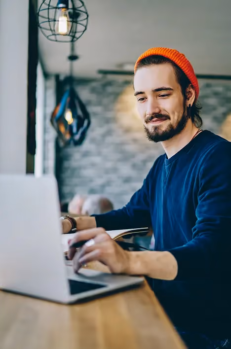 Man wearing an orange beanie and navy sweater working on a laptop at a wooden table in a modern room.
