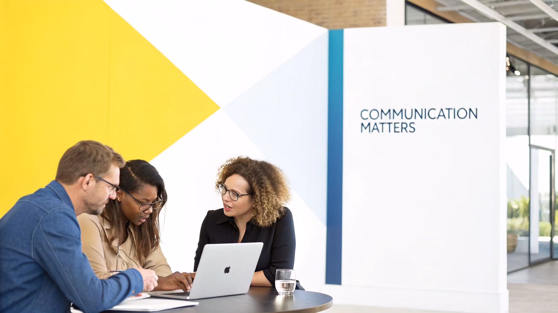 Three colleagues discussing work around a laptop at a round table in an office with 'Communication Matters' on the wall.