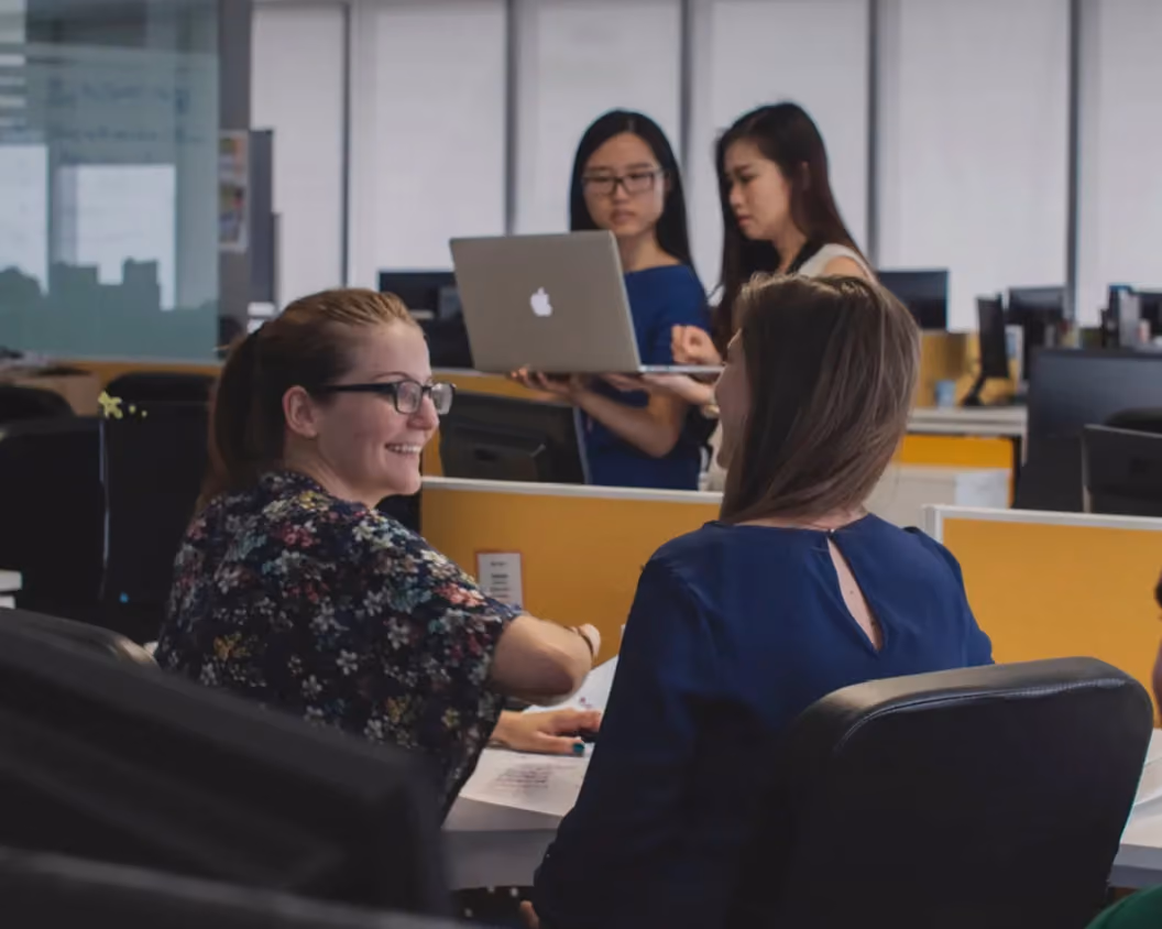 Four women collaborating in an office; two seated talking and two standing using a laptop.