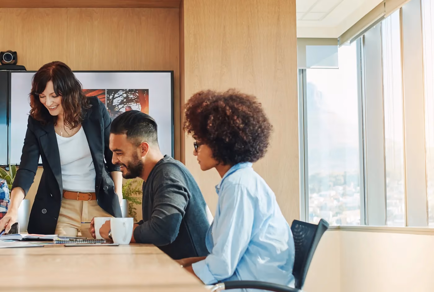 Three coworkers in a meeting room, one standing and pointing at documents, two seated and engaged.