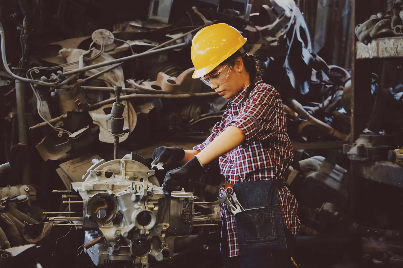 Woman in a yellow hard hat and plaid shirt working on machinery in a cluttered workshop.