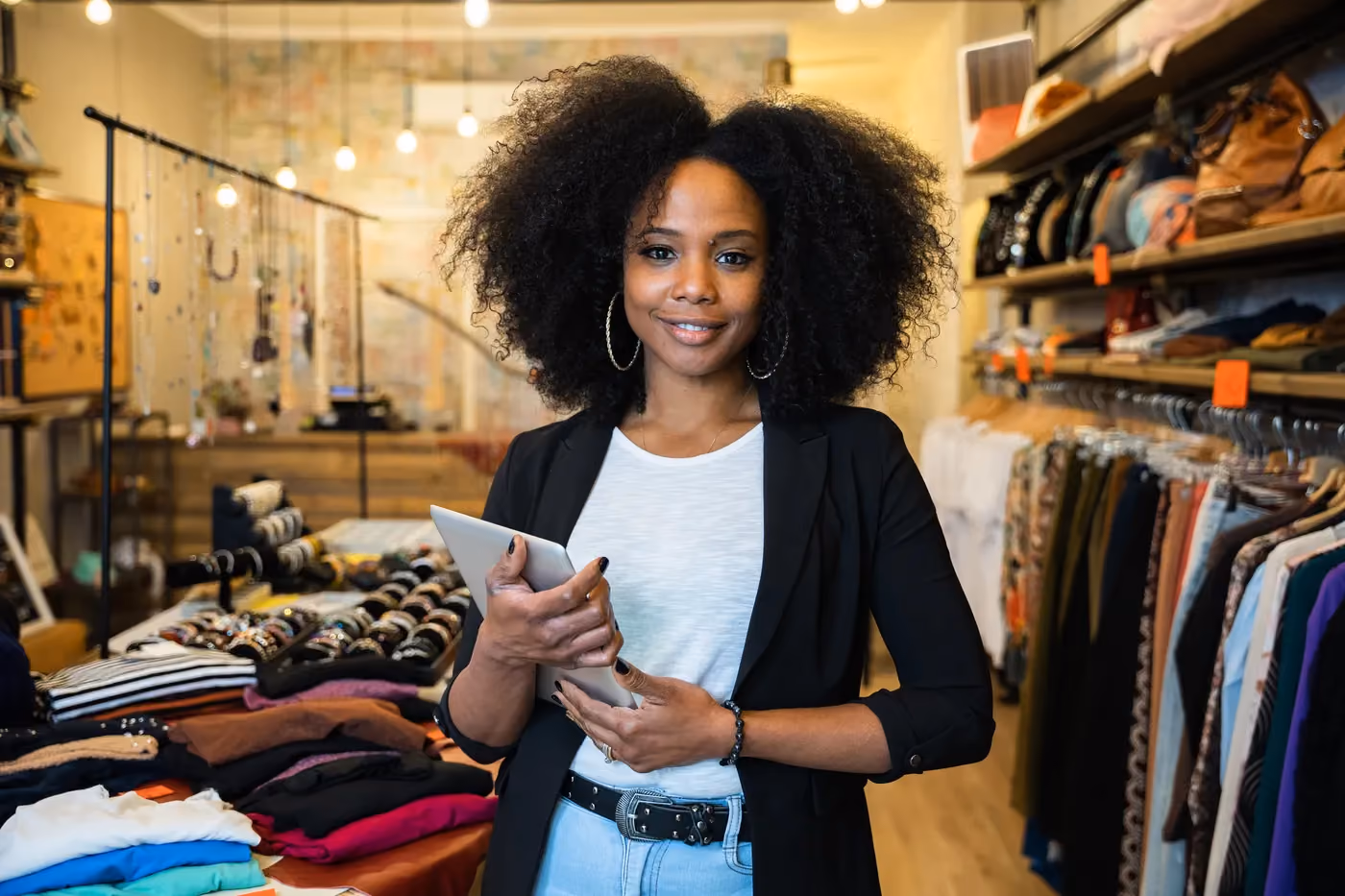 Smiling woman with curly hair holding tablet in a boutique clothing and accessories store.