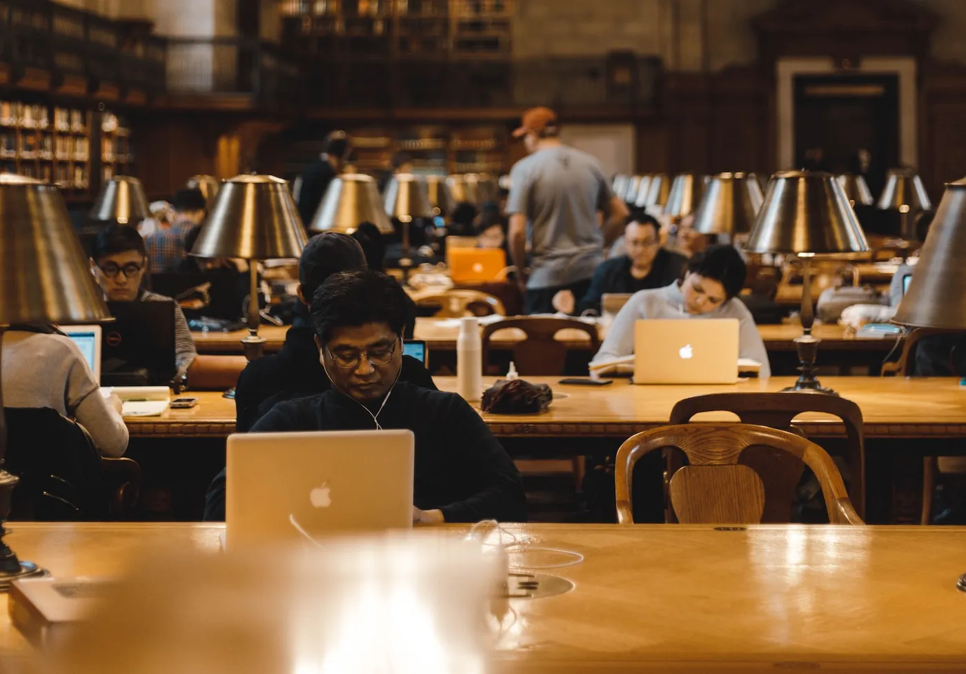 People studying on laptops at long wooden tables with desk lamps in a large library reading room.