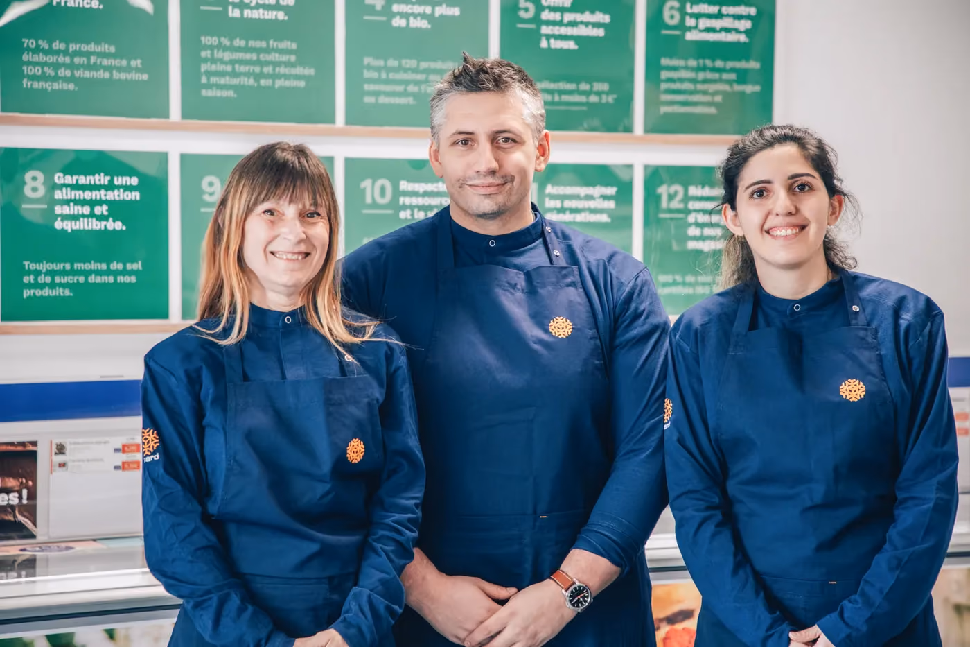 Three smiling food service workers wearing navy aprons standing in front of green informational posters.