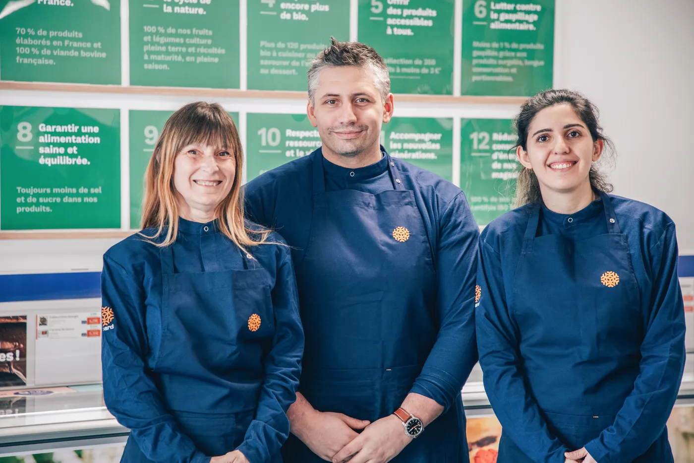 Three smiling food service workers wearing navy aprons standing in front of green informational posters.