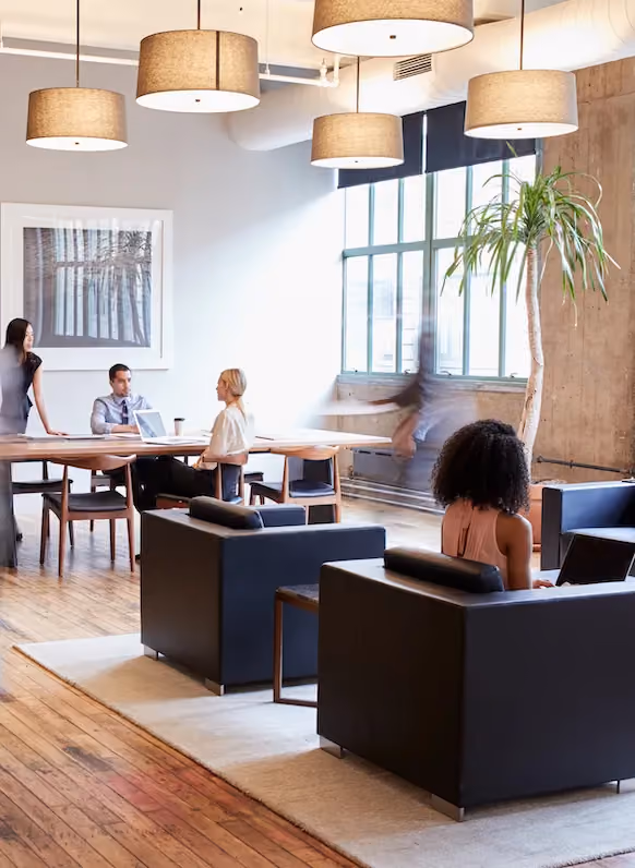 Modern office with a group meeting around a table and one woman sitting on a black chair using a laptop.