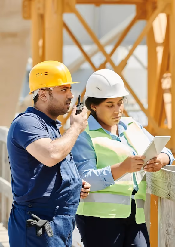Two construction workers with helmets, one using a walkie-talkie, the other checking a tablet.