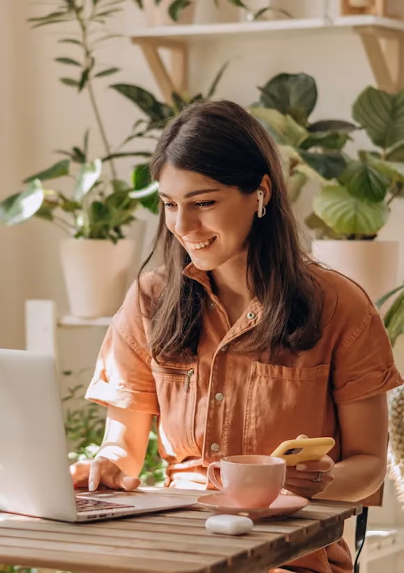 Smiling woman with earbuds uses laptop and holds smartphone at wooden table with coffee cup.