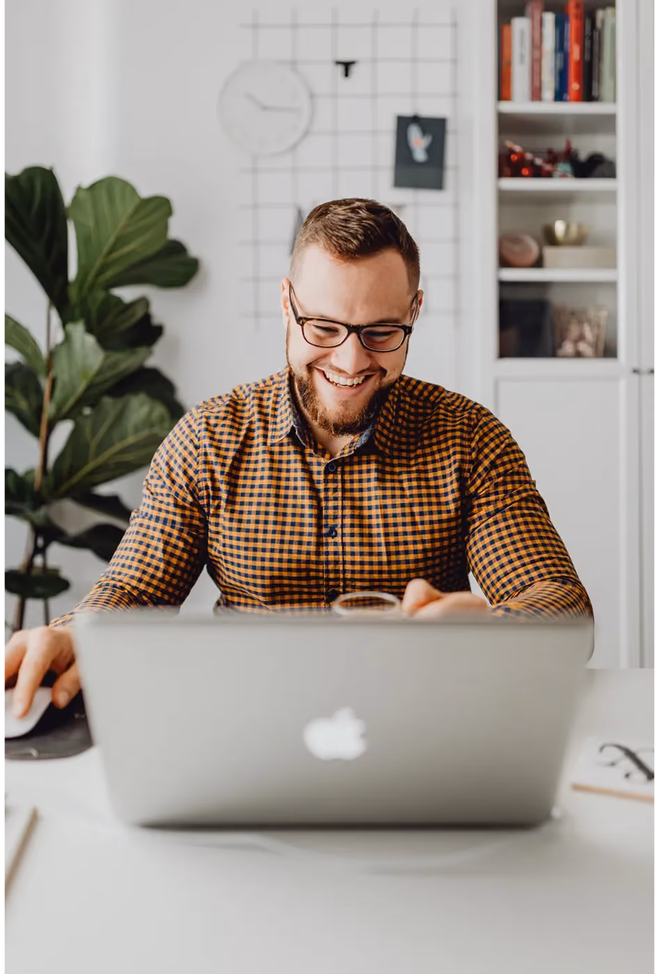 Smiling man in glasses and checkered shirt working on a laptop at a bright home office desk.