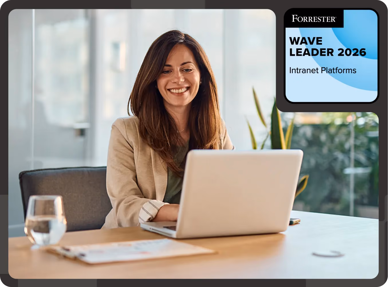 Smiling woman in tan blazer using a laptop at desk with glass of water and documents nearby.