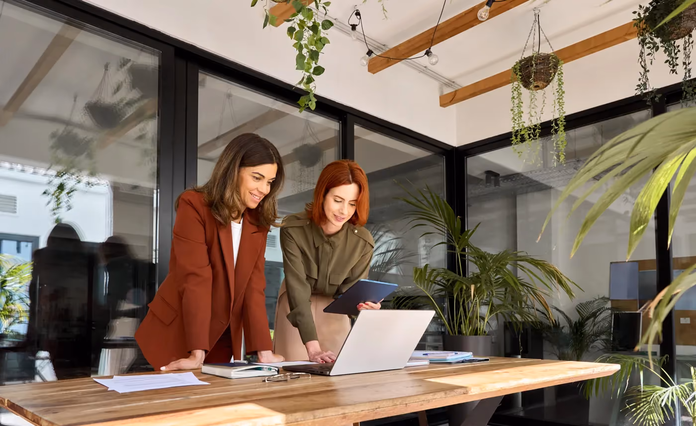Two women standing at a wooden table working on a laptop in a plant-filled modern office.