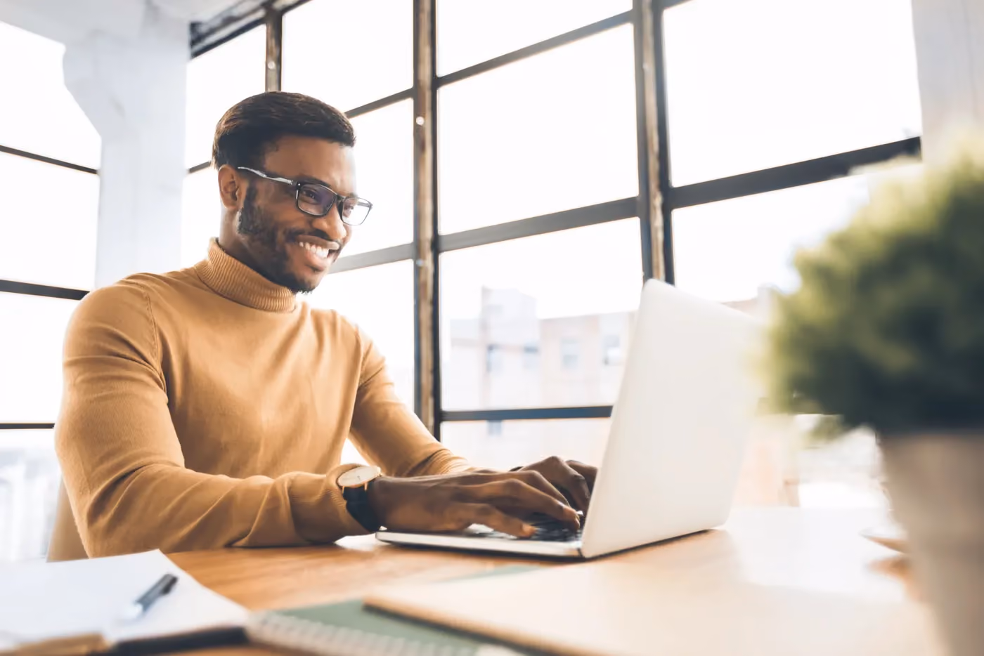 Smiling man in glasses and brown sweater working on laptop at a bright office desk.
