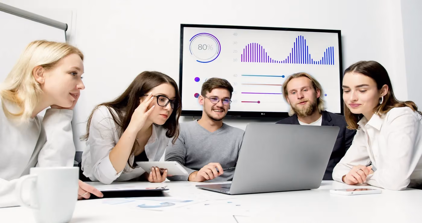 Five young professionals sitting around a desk with a laptop and discussing data charts on a screen.