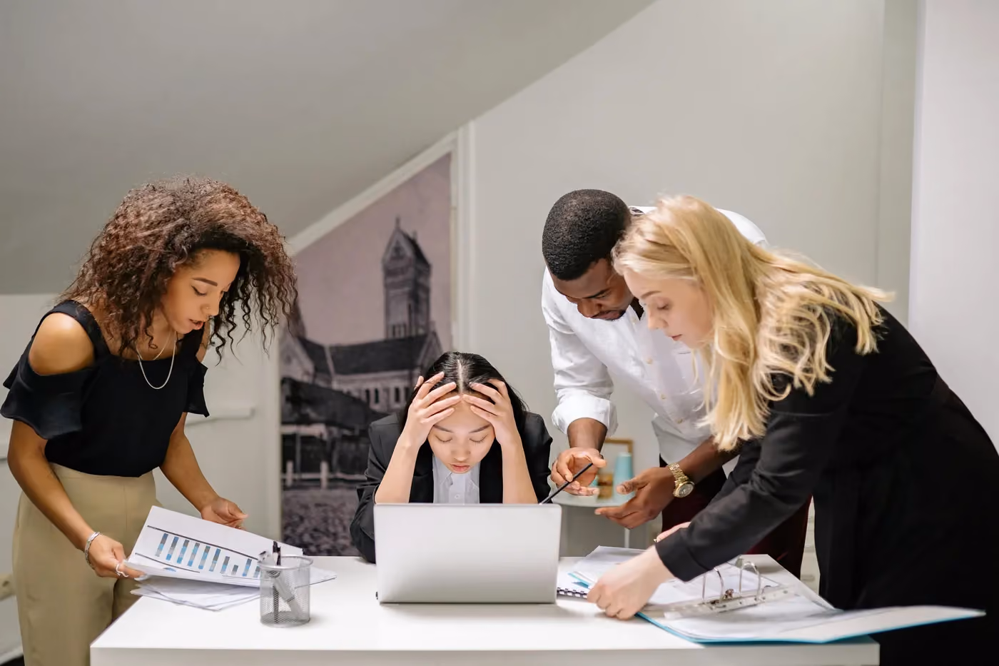 Four diverse coworkers gathered around a laptop, one stressed, others discussing documents and charts.