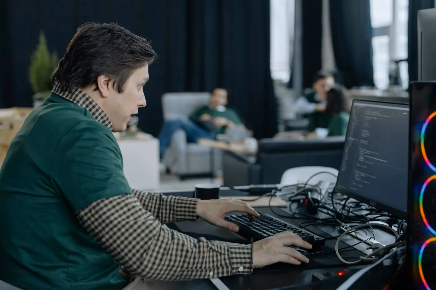 Man focused on coding at a computer in a modern office with other people in the background.