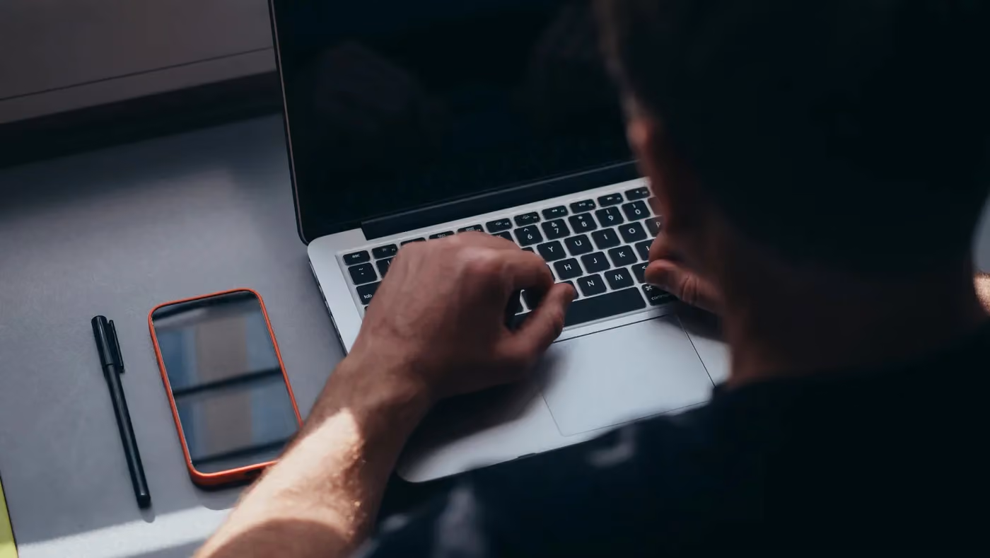 Person typing on a laptop keyboard next to a smartphone and black pen on a gray desk.
