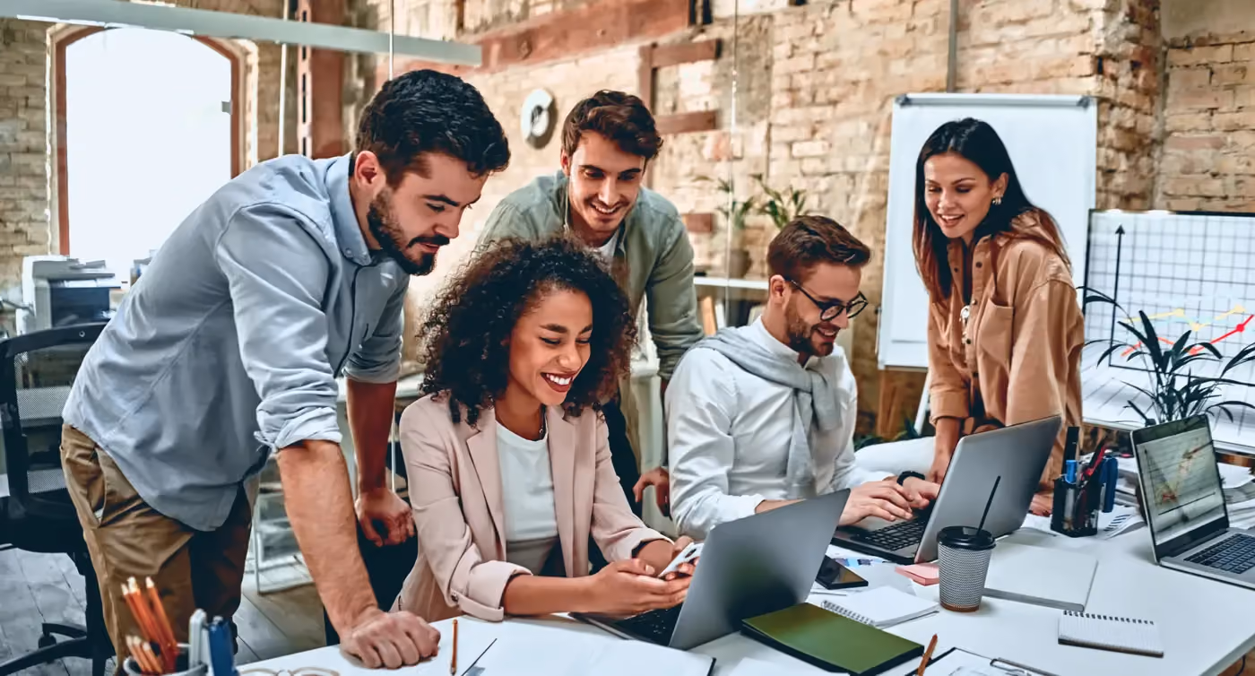 Five diverse coworkers smiling and collaborating at a laptop and smartphone in a modern office.