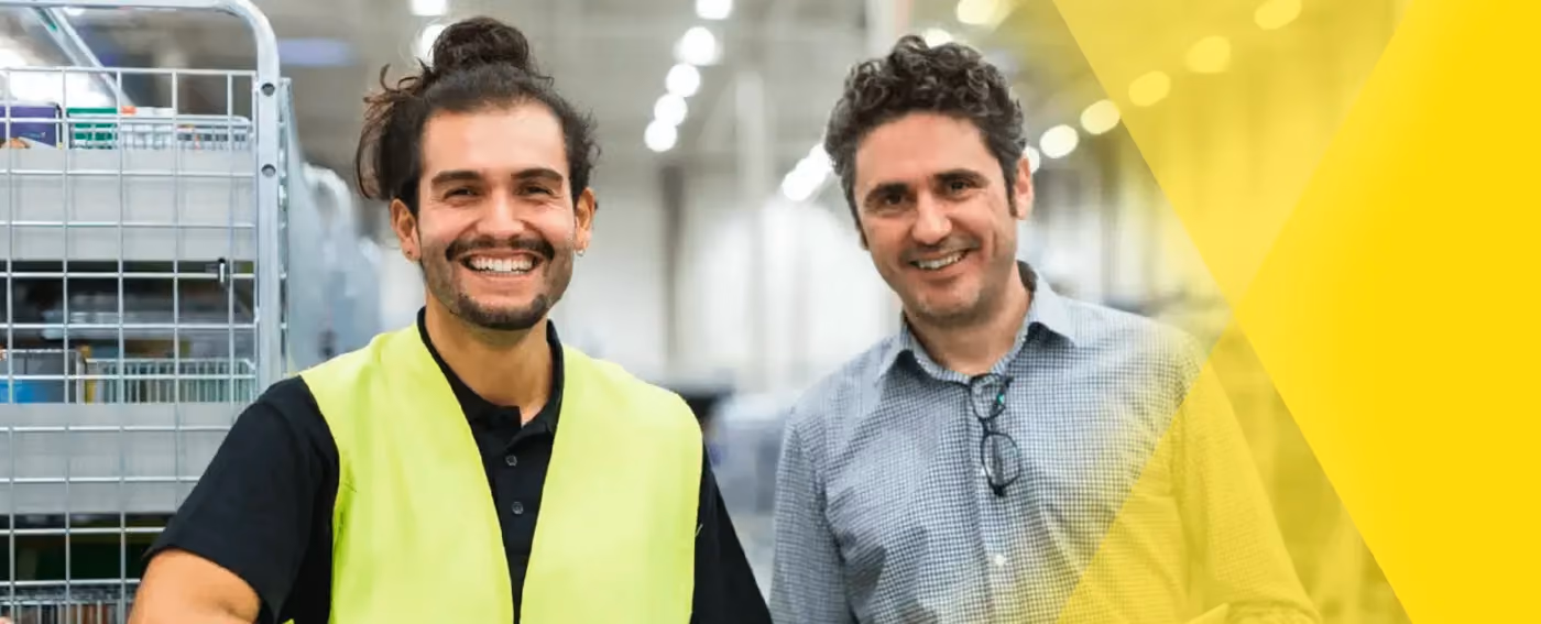 Smiling warehouse worker in yellow vest and man in checked shirt standing together inside a facility.