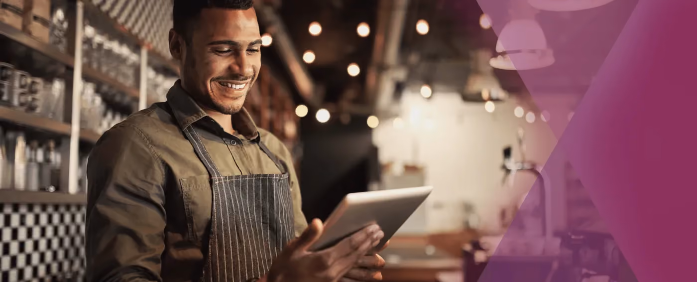Smiling barista wearing apron looking at a tablet in a cozy cafe with warm lighting.