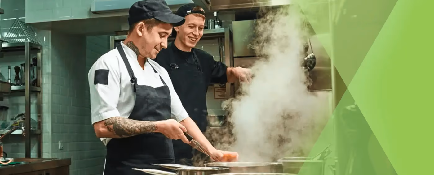 Two chefs smiling and cooking with steam rising from pots in a commercial kitchen.