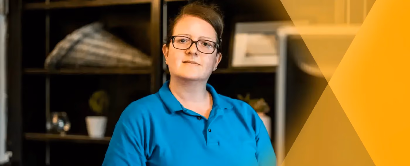 Serious woman wearing glasses and blue polo shirt standing indoors with shelves in background.