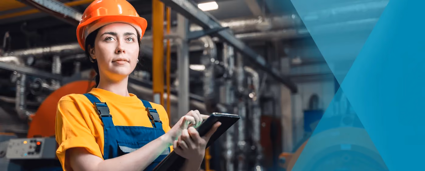 Female factory worker wearing orange helmet and overalls using a digital tablet in a manufacturing plant.