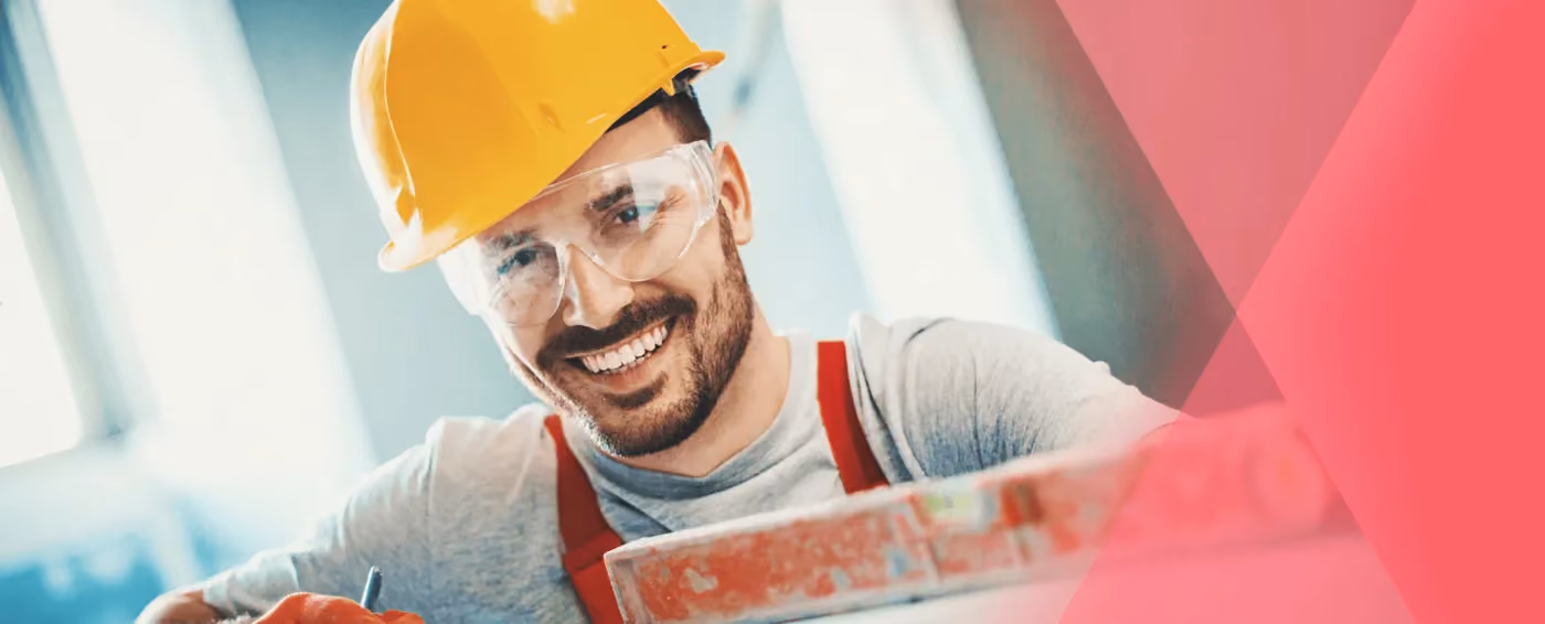 Smiling male construction worker wearing a yellow hard hat and safety glasses indoors.