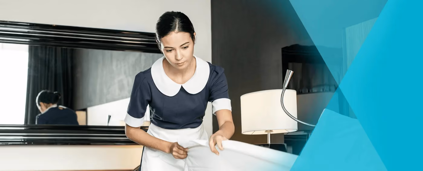 Hotel housekeeper in uniform making a bed with white sheets in a guest room.