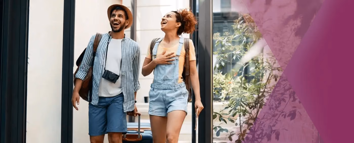 Smiling man and woman with backpacks walking with luggage outside a building on a sunny day.