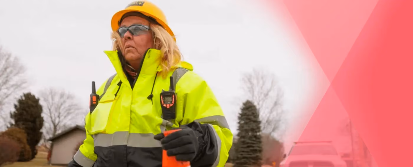 Woman in safety gear holding a traffic cone outdoors with trees and building in background.