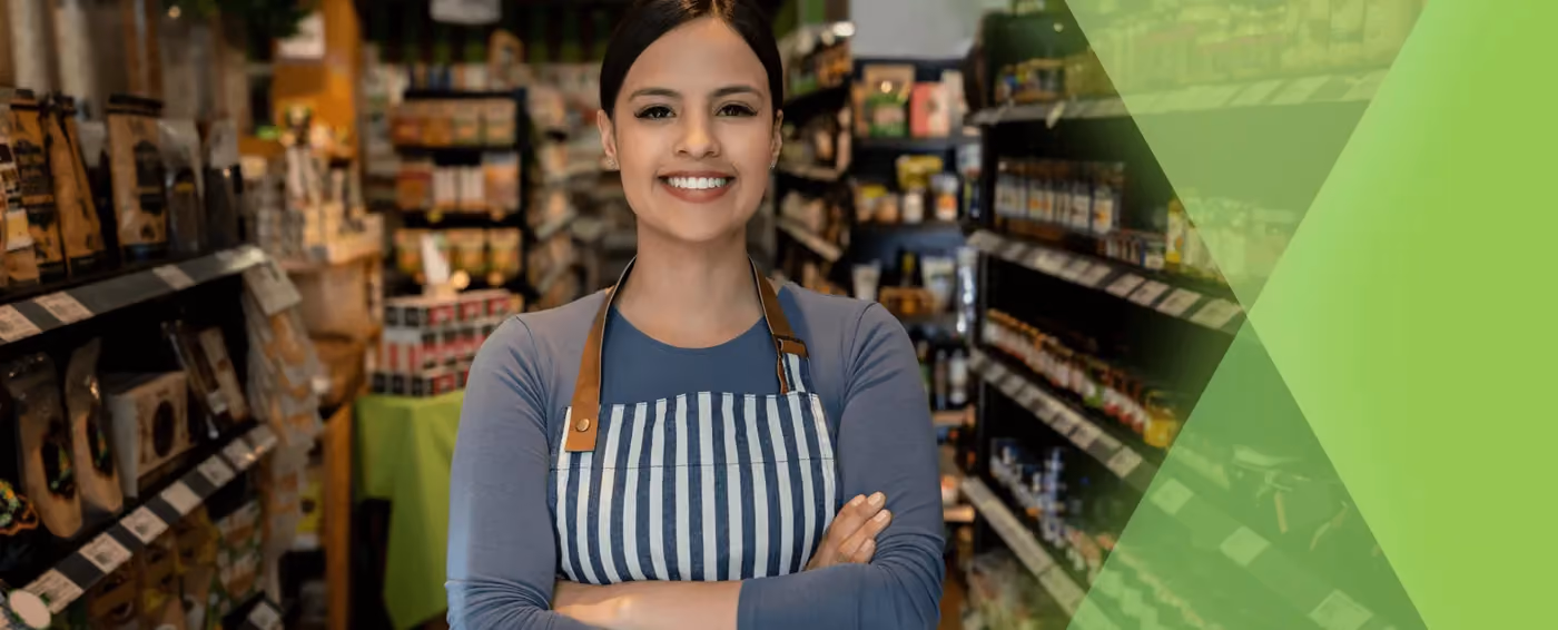 Smiling woman in a striped apron standing with arms crossed in a store aisle with shelves of products.