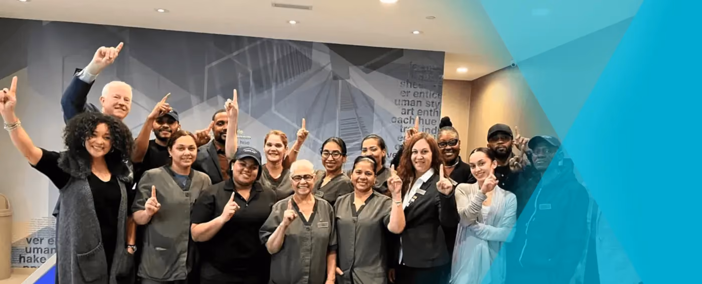 Group of diverse healthcare workers and staff smiling and holding up one finger in a lobby area.