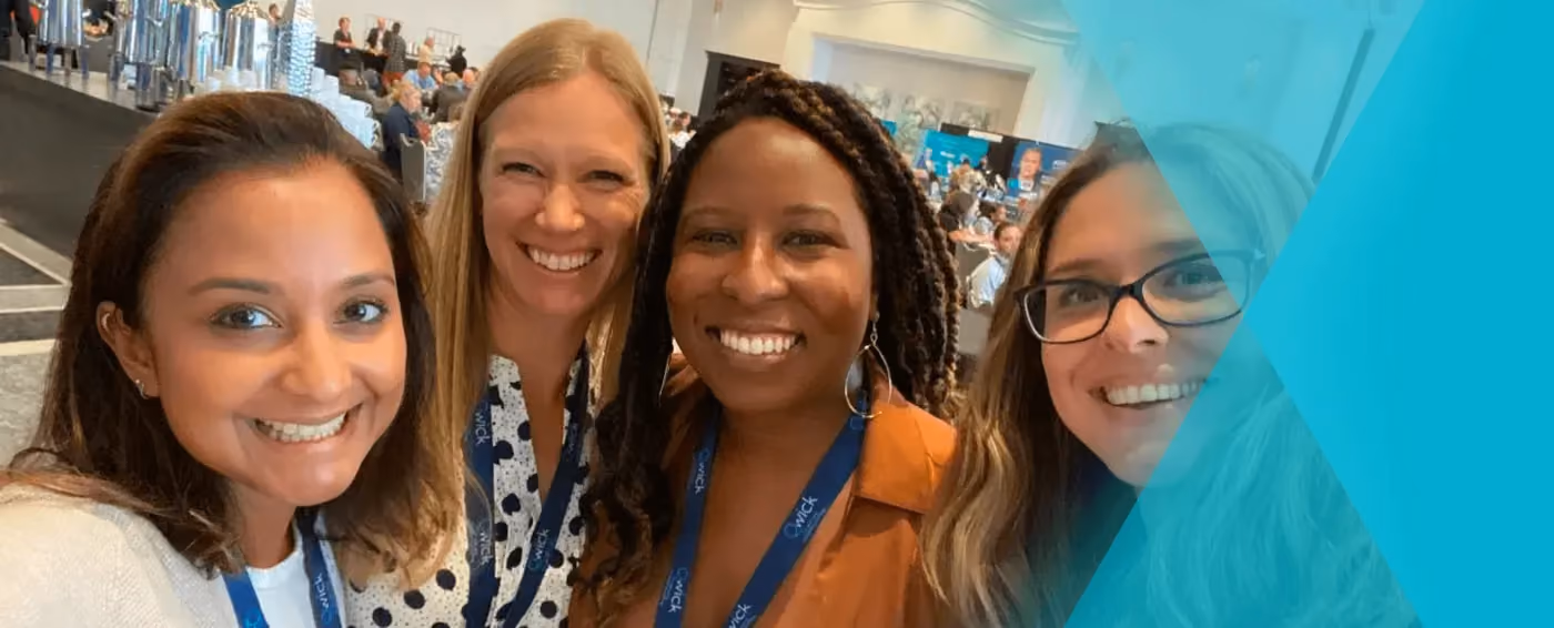Four smiling women wearing conference lanyards posing together indoors at a crowded event.