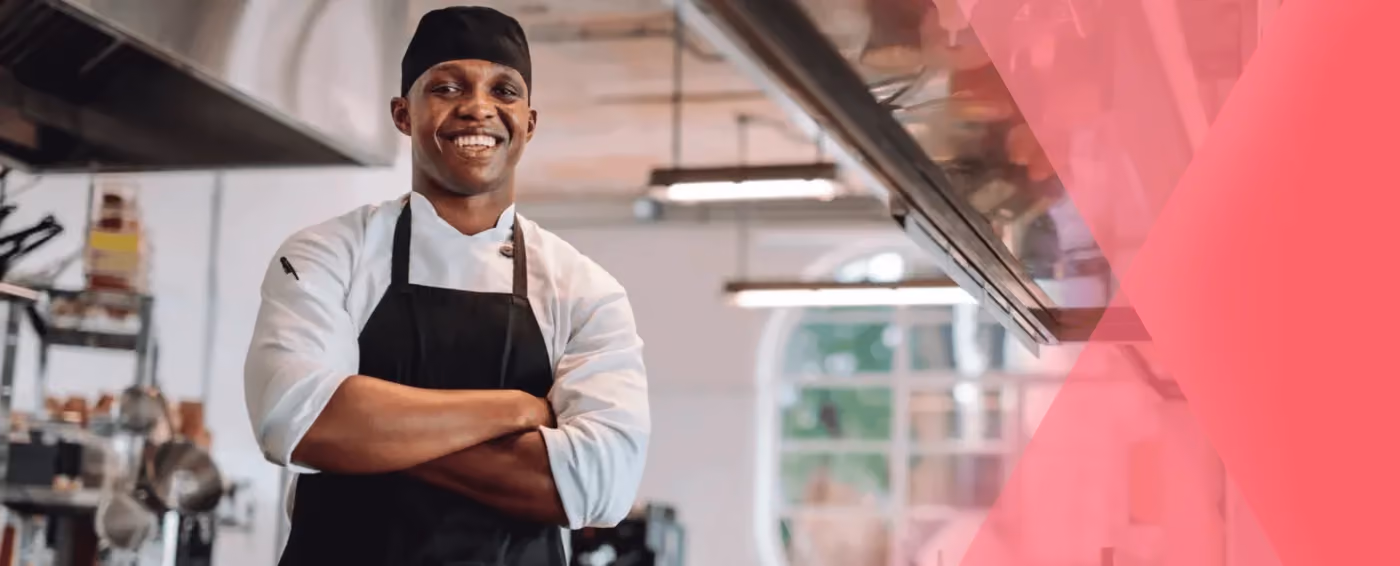 Smiling chef in a black apron and hat standing with arms crossed in a professional kitchen.
