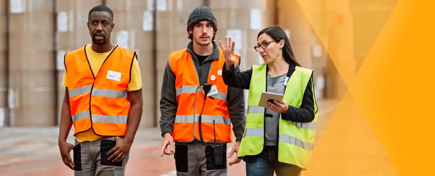Two men in orange safety vests and a woman in a yellow vest holding a tablet in a warehouse talk.