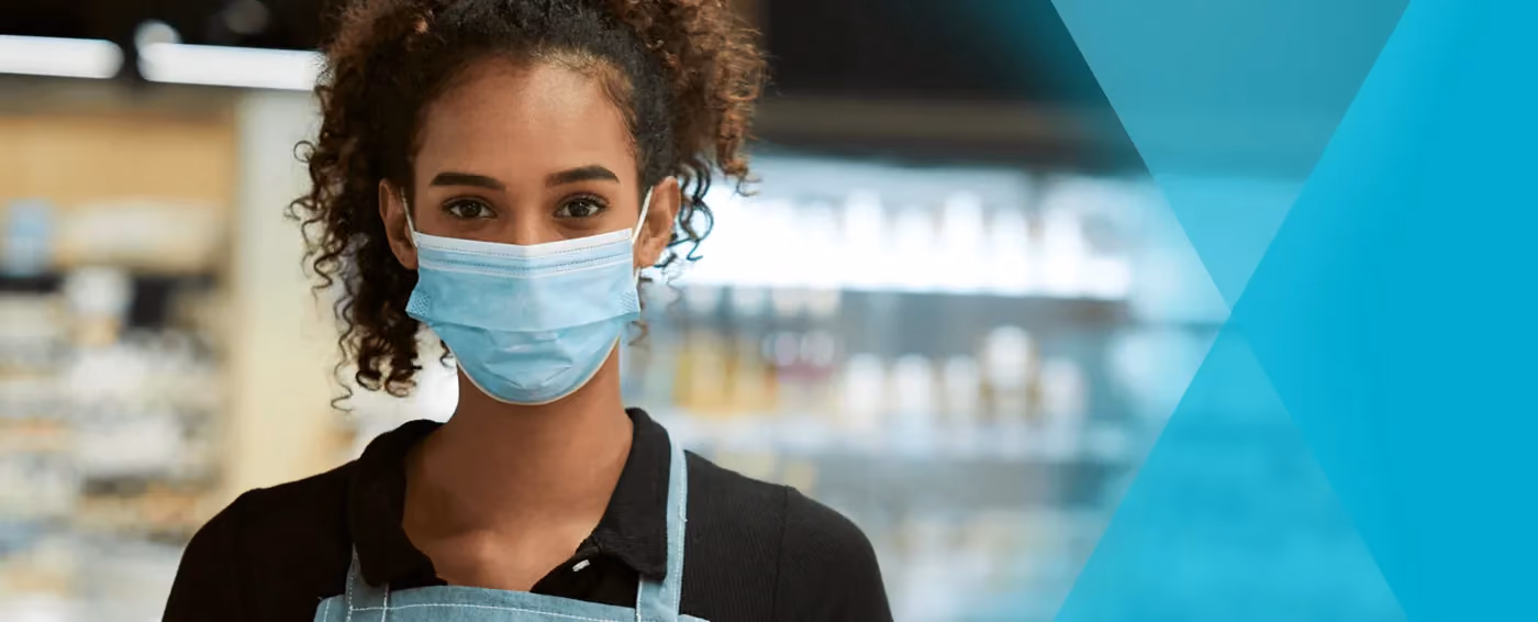 Young woman with curly hair wearing medical mask and apron stands in a grocery store aisle.