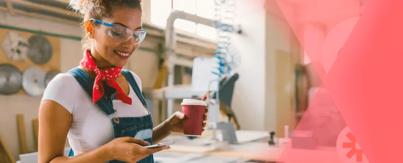 Smiling woman in safety goggles and overalls holding a coffee cup and using a smartphone in a workshop.