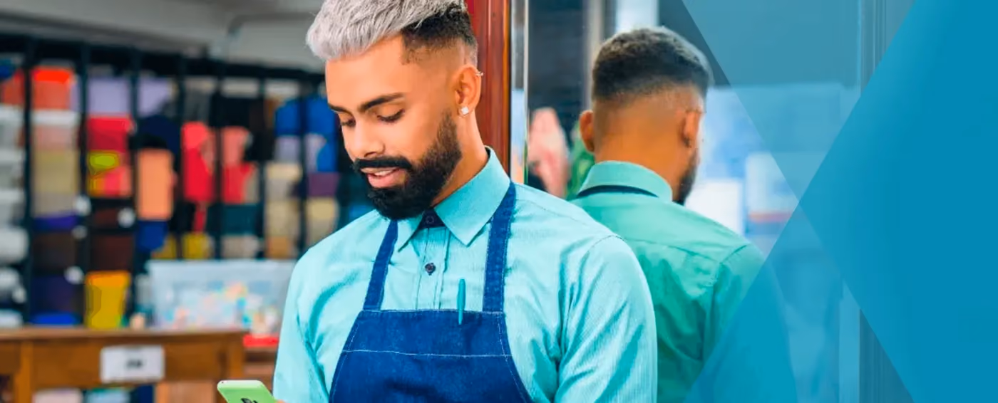 Man with gray hair and beard wearing apron, looking at phone with colorful fabric rolls in background.