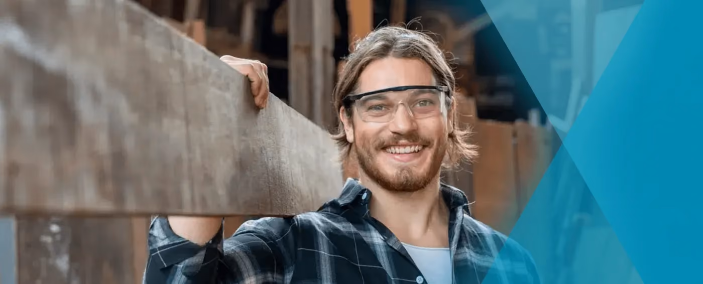 Smiling man wearing safety glasses and plaid shirt holding a wooden plank in a workshop.