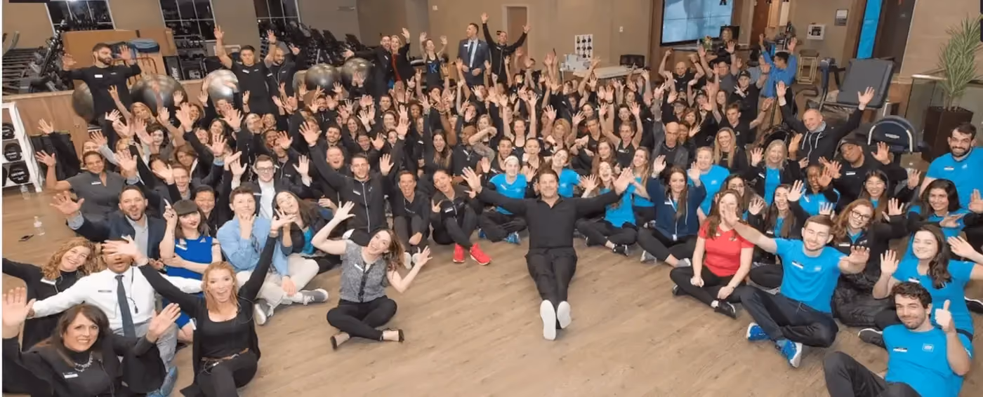 Large group of people sitting on a floor in a gym, smiling and waving at the camera.