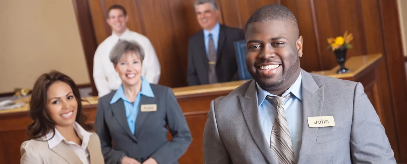 Smiling hotel staff in business attire, with man named John in front at reception desk.