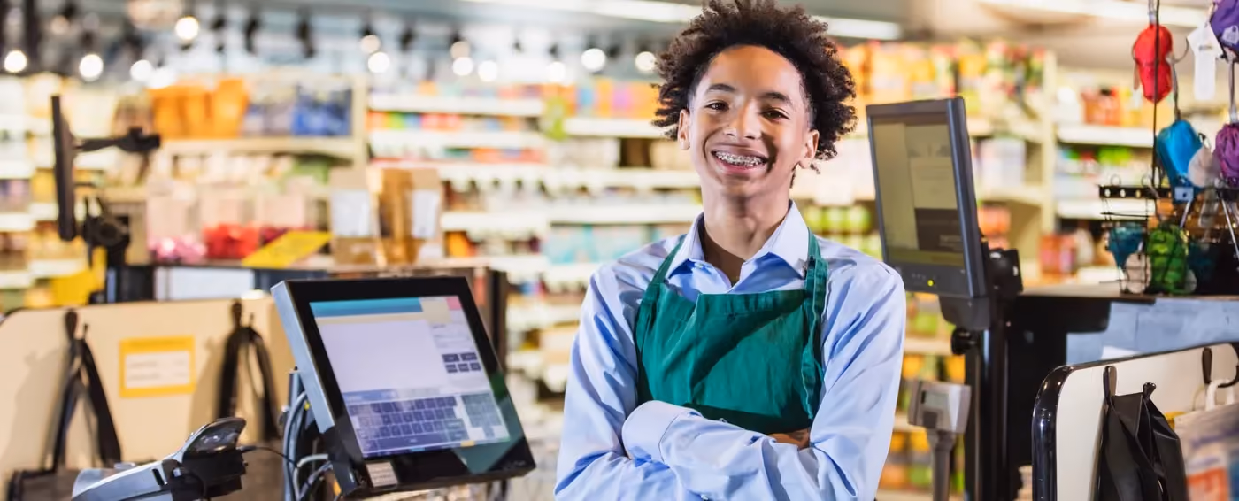 Smiling young male cashier wearing green apron standing at checkout with crossed arms in a store.